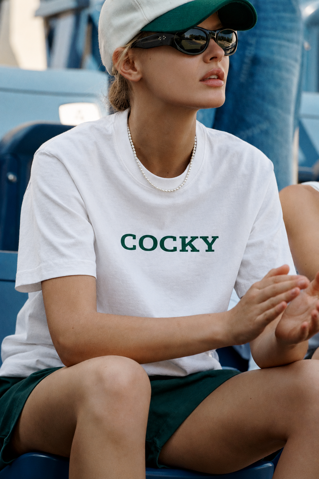 White COCKY T-shirt styled with green shorts, cap, sunglasses and pearls in stadium seats